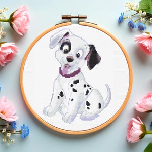 May include: A cross-stitch embroidery of a Dalmatian puppy in a wooden hoop. The puppy is white with black spots, wearing a purple collar. The background is white, and the hoop is surrounded by pink and blue flowers.
