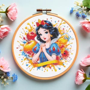 Puede incluir: Bordado de punto de cruz de Blancanieves en un aro de madera. El diseño presenta a Blancanieves con un vestido azul y un lazo rojo, rodeada de salpicaduras de pintura coloridas y manzanas. El bordado está sobre un fondo azul claro con flores rosas y azules.