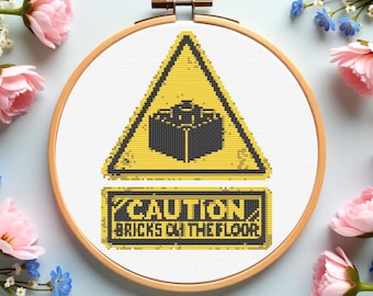 May include: A cross-stitch embroidery in a wooden hoop features a yellow caution sign with a black brick design and the text "CAUTION BRICKS ON THE FLOOR". The design is surrounded by pink and blue flowers.