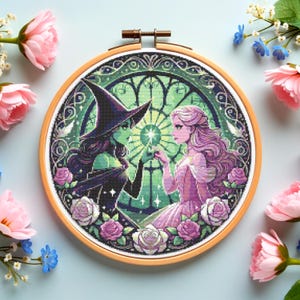 Op de afbeelding: Kruissteekborduurwerk in een houten hoepel met twee figuren: een groenhuidige heks in een zwarte hoed en jurk en een vrouw in een roze jurk. Het ontwerp bevat een glas-in-lood-effect en paarse rozen.