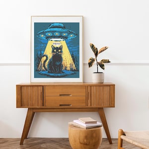 May include: Framed pixel art print featuring a black cat illuminated by a UFO's beam of light. The artwork includes the word "MEOW?" above the cat. The print is displayed on a wooden console table with a plant and a small side table.