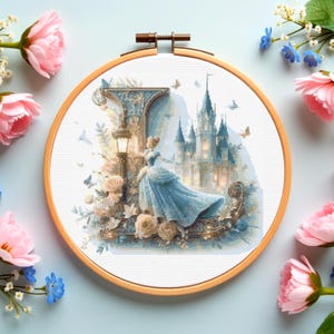 May include: A cross-stitch embroidery hoop featuring a fairytale scene. The design depicts a woman in a blue gown, a castle, and floral elements. The wooden hoop is surrounded by pink and blue flowers on a light blue background.