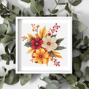 May include: A framed cross-stitch floral design featuring a bouquet of flowers in shades of red, orange, and cream, with green leaves and berries. The artwork is set against a white background within a white frame.