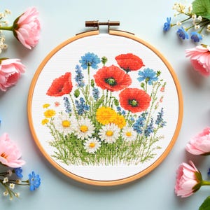 May include: A completed cross-stitch embroidery of a wildflower bouquet in a wooden hoop. The design features red poppies, blue cornflowers, yellow dandelions, and white daisies. The hoop is surrounded by pink and blue flowers on a light blue background.