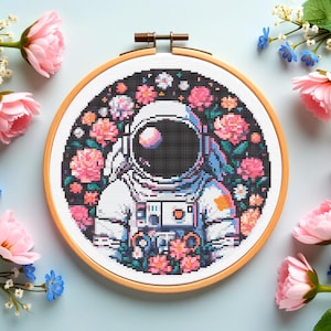 May include: A cross-stitch embroidery hoop featuring an astronaut surrounded by pink and white flowers against a black background. The astronaut is wearing a white spacesuit with blue and orange accents. The wooden hoop is round.
