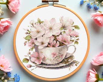 Puede incluir: Un bordado de punto de cruz de una taza de té rebosante de flores rosas y blancas, en un aro de madera. La taza y el platillo están decorados con motivos florales. Flores rosas y azules dispuestas alrededor del aro sobre un fondo azul claro.