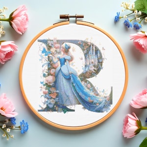 May include: A cross-stitch embroidery in a wooden hoop features a princess in a blue gown within a floral-decorated letter "R". The design includes a castle, butterflies, and pastel flowers. Pink and blue flowers surround the hoop.