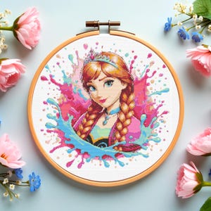Puede incluir: Un bastidor de bordado de punto de cruz con un retrato de una princesa con trenzas rojizas, una tiara y un diseño de salpicaduras rosas y azules. El bordado está en un bastidor de madera, rodeado de flores rosas y azules.