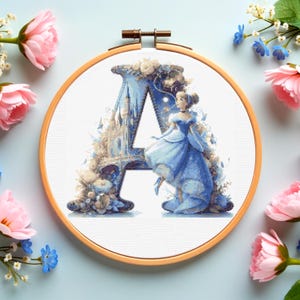 May include: A cross-stitch embroidery hoop with the letter "A" in a Cinderella design. The letter is blue and white, with a castle and Cinderella in a blue gown. The hoop is surrounded by pink and blue flowers.