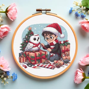 May include: A cross-stitch hoop depicting a Christmas scene with a boy in a Santa hat and scarf, alongside a white owl. A Christmas tree and wrapped gifts are also visible. The hoop is framed by pink and blue flowers.