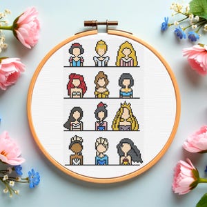 May include: A cross-stitch embroidery hoop featuring pixelated images of various princesses. The princesses are depicted with distinct hairstyles and outfits, showcasing a range of colors. The wooden hoop is surrounded by pink and blue flowers, creating a decorative display.