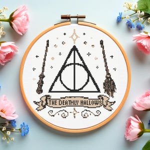 May include: A circular wooden embroidery hoop containing a cross-stitch pattern on white fabric. The design features a black Deathly Hallows symbol, a wand, a broomstick, and a banner with the text THE DEATHLY HALLOWS in black lettering.