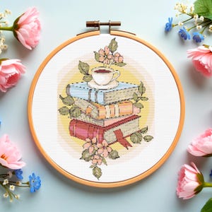 Puede incluir: Bordado de punto de cruz en un aro de madera que muestra una pila de libros con una taza de té encima. Los libros son de color azul, naranja y rojo, con hojas verdes y flores rosas. El fondo es un degradado amarillo suave.