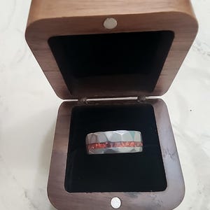 May include: A silver ring with a geometric design and a band of orange stones, displayed in an open wooden box with a black velvet interior. The box is made of dark wood and has a magnetic closure.