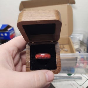 May include: A red and black ring is presented in an open wooden ring box. The ring features a speckled design. The box is crafted from light brown wood and has a dark velvet lining.