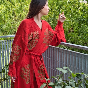 Op de afbeelding: Een felrode kimono met goudkleurige bladvormige patronen. De kimono is in de taille vastgebonden met een bijpassende riem. De mouwen zijn lang en de stof lijkt licht. De achtergrond toont weelderig groen gebladerte en een metalen reling.