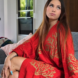 Op de afbeelding: Een rode robe met goudkleurige koraalachtige borduursels. De robe heeft lange mouwen en een gefranjerde zoom. De persoon zit op een grijze bank.