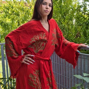 Op de afbeelding: Een felrode kimono met goudkleurige geborduurde boompatronen. De kimono heeft wijde mouwen met gefranjerde randen en een bijpassende ceintuur. Het kledingstuk is gemaakt van een lichte stof, perfect om in te loungen of als strandcover-up.