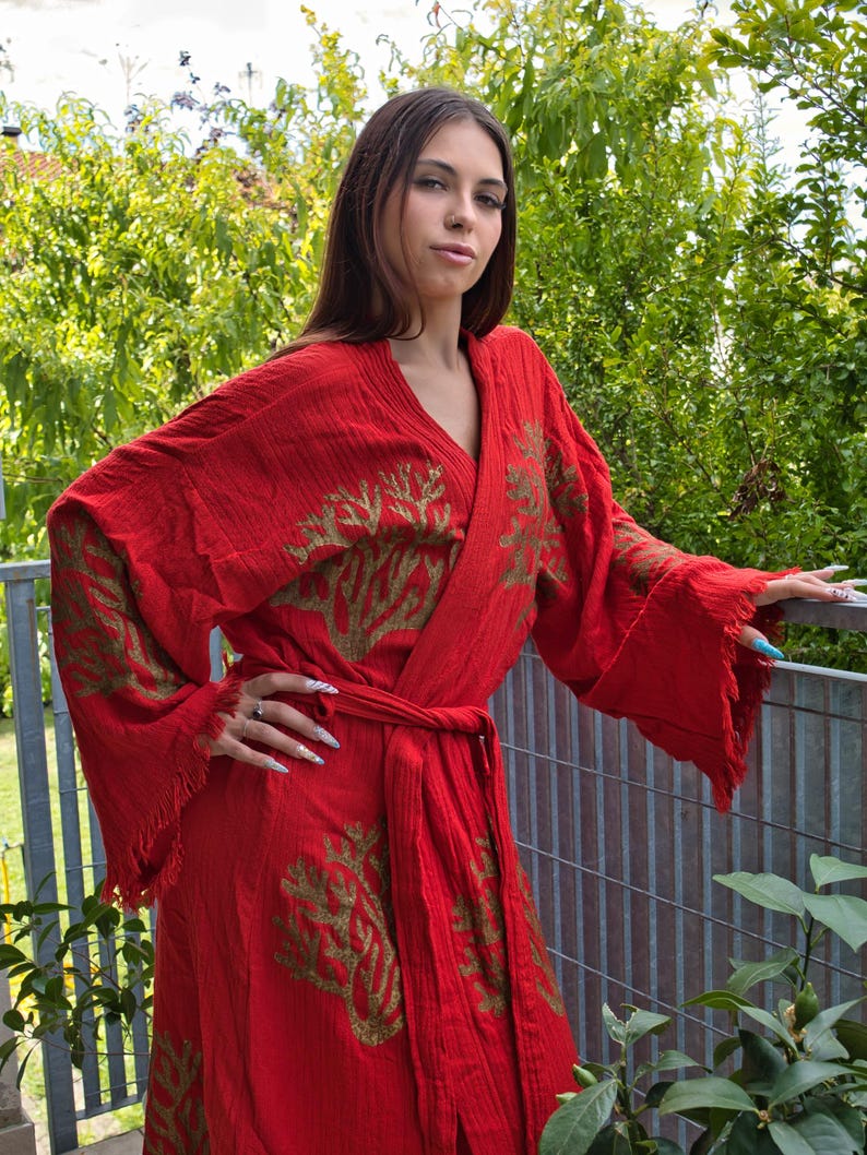 Op de afbeelding: Een felrode kimono met goudkleurige geborduurde boompatronen. De kimono heeft wijde mouwen met gefranjerde randen en een bijpassende ceintuur. Het kledingstuk is gemaakt van een lichte stof, perfect om in te loungen of als strandcover-up.