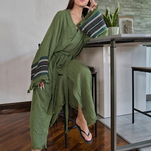 Può includere: Accappatoio stile kimono verde oliva e pantaloni abbinati con bordi sfilacciati. L'accappatoio presenta fasce decorative di cuciture nere, bianche e verde acqua sulle maniche. L'outfit è abbinato a infradito nere.