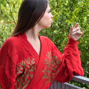Op de afbeelding: Een rode kimono-achtige robe met goud geborduurde bloemmotieven. De robe heeft een V-hals en lange mouwen. De mouwen hebben een gerafelde rand. De achtergrond is weelderig groen gebladerte.