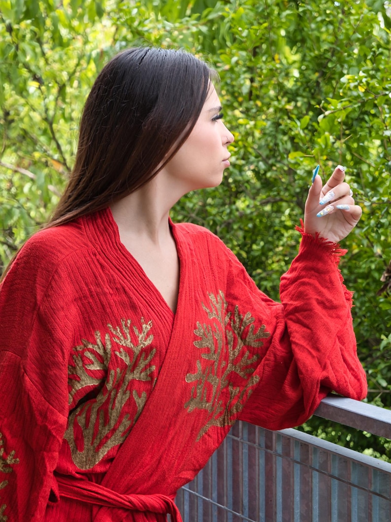 Op de afbeelding: Een rode kimono-achtige robe met goud geborduurde bloemmotieven. De robe heeft een V-hals en lange mouwen. De mouwen hebben een gerafelde rand. De achtergrond is weelderig groen gebladerte.