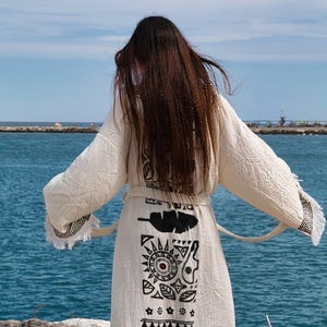 May include: A cream-colored robe with long, wide sleeves and a tie waist. The back features a vertical panel of black printed designs, including a feather, a sun, geometric shapes, and leaf motifs, with fringed edges on the sleeve cuffs.