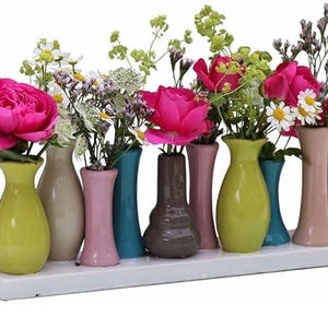 Könnte beinhalten: Eine Anordnung kleiner, farbenfroher Keramikvasen, jede mit frischen Blumen. Die Vasen sind in verschiedenen Farben gehalten, darunter Gelb, Rosa, Türkis und Braun. Die Blumen sind eine Mischung aus rosa Rosen, Gänseblümchen und Grün, angeordnet auf einer weißen rechteckigen Basis.