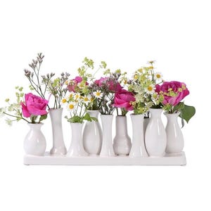 May include: A white ceramic bud vase set holding a variety of fresh flowers. The flowers include pink roses, white daisies, and other greenery. The vases are arranged on a rectangular base, creating a decorative floral display.