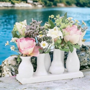 May include: A collection of white ceramic bud vases holding an assortment of fresh flowers. The flowers include pink roses, white daisies, and other greenery. The vases are arranged on a white rectangular tray, with a blurred water background.