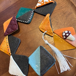 May include: A handmade, colorful necklace featuring diamond-shaped pendants. The pendants are decorated with various designs, including a mushroom, ghosts, and floral patterns. The necklace has a white tassel and is set on a wooden surface.