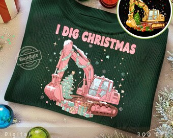 Puede incluir: Sudadera verde oscuro con el texto "I DIG CHRISTMAS" y una excavadora rosa decorada con un árbol de Navidad y guirnaldas. El diseño incluye copos de nieve y cajas de regalo, con un gráfico circular de una excavadora amarilla.