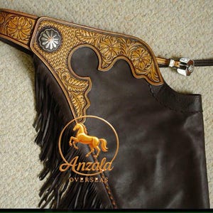 May include: Dark brown leather chaps with tan and gold floral and geometric tooling. The chaps have a silver buckle, side fringe, and the "Anzala Overseas" logo with a golden horse. The chaps are likely for riding.