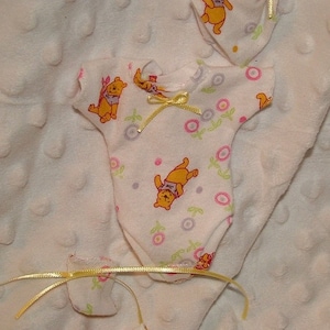 May include: A white baby onesie, hat, and two small accessories, all decorated with a yellow bear cartoon character and pink floral accents. The onesie has a yellow bow at the neckline. The items are on a white, textured surface.
