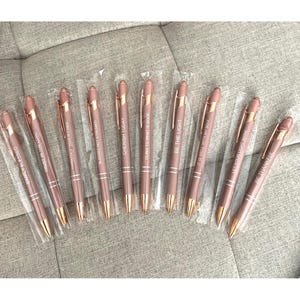 May include: A collection of rose gold pens with inspirational quotes in clear plastic packaging. The pens have gold accents and are engraved with phrases like "Simply Blessed," "Be the Light," and "Cherish Today." The pens are displayed on a gray surface.