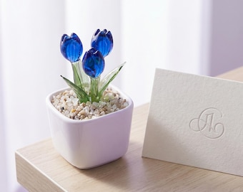 Pode incluir: Um pequeno vaso quadrado de cerâmica branca contém três flores de tulipa de vidro azul com caules e folhas verdes. O vaso é preenchido com pequenas pedras claras. À direita, há um cartão branco com um desenho em relevo.
