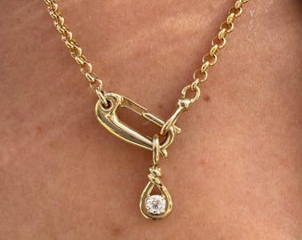 Puede incluir: Collar dorado con un colgante de cierre grande y un dije en forma de lágrima. El dije contiene una sola piedra preciosa redonda y transparente. La cadena es de diseño de eslabones y el collar se usa alrededor del cuello.