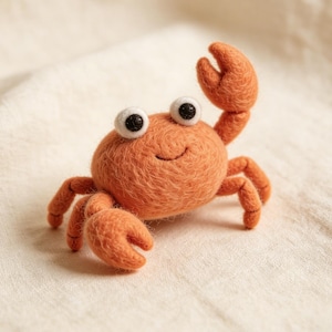 Peut inclure: Un petit crabe en feutre orange avec des yeux globuleux et un visage souriant. Le crabe a deux grandes pinces et plusieurs pattes. Le crabe est posé sur un fond texturé de couleur crème.