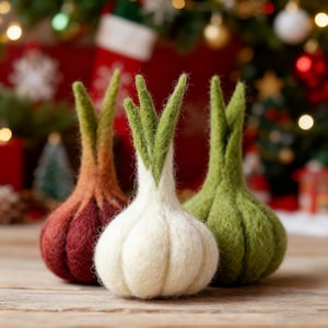 May include: Three felted onion-shaped ornaments in shades of red, white, and green. Each ornament has a bulbous body and green, sprout-like tops. The ornaments are arranged on a wooden surface with a blurred Christmas tree background.
