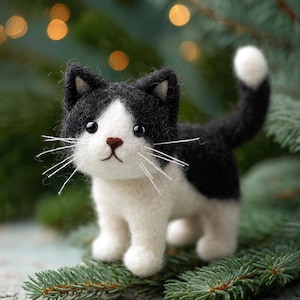 May include: A handcrafted felt cat ornament in black and white. The cat features a white face, chest, and paws, contrasted by black patches on its head and back. It has a small brown nose, black eyes, and white whiskers. The cat is posed on a green surface.