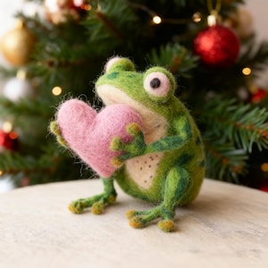 Könnte beinhalten: Eine handgefertigte grüne Froschfigur, die ein rosa Herz hält. Der Frosch hat große Augen und gelbe Füße. Im Hintergrund befindet sich ein Weihnachtsbaum mit roten und goldenen Kugeln und Lichterketten.