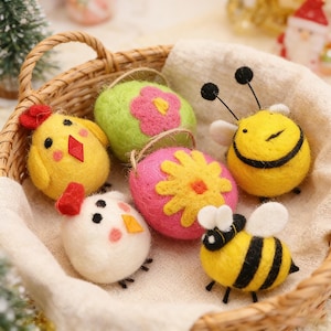 Op de afbeelding: Een rieten mand bevat zes vilten paasversieringen. De versieringen omvatten twee gele kuikens, twee bijen en twee eivormige ornamenten versierd met bloemen. De ornamenten zijn gemaakt van vilt en hebben lussen om op te hangen.