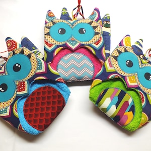 May include: Three colorful owl-shaped kitchen towels with different patterned designs. Each towel has a blue eye with a black pupil and a bright colored towel insert.
