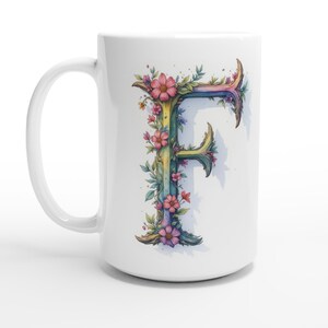 Può includere: Tazza in ceramica bianca con un disegno ad acquerello della lettera "F", ornata di fiori rosa e foglie verdi. La lettera presenta una sfumatura di colori, tra cui giallo, blu e viola. La tazza ha un manico curvo.