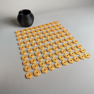 May include: A black, matte-finish ceramic vase sits next to a collection of small, gold-colored, hexagonal objects. Each object has a square center hole. The objects are arranged in a geometric pattern.