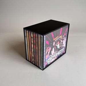 May include: A black CD storage box filled with multiple CDs. The front CD cover features the word "Spaghetti" in a pink and white design with a photo of a group of people. The other CD covers are visible on the side.