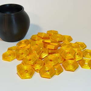 May include: A black, round pot sits next to a pile of translucent, golden-yellow, hexagonal tokens. The tokens have a geometric design with a square hole in the center. The pot and tokens are on a white surface.