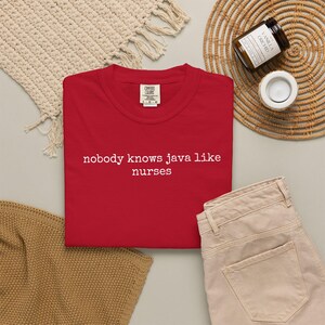 Puede incluir: Una camiseta roja con el texto blanco "nobody knows java like nurses". La camiseta se muestra con pantalones beige, un salvamanteles tejido y una vela.