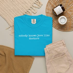 Puede incluir: Una camiseta turquesa Comfort Colors con el texto "nobody knows java like doctors" en blanco. La camiseta se muestra con pantalones beige, un mantel individual tejido y una vela de vainilla orquídea.