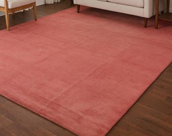 Puede incluir: Una alfombra rectangular grande de color coral domina una escena de sala de estar. La alfombra se asienta sobre un piso de madera oscura, con un sofá beige, un sillón y una planta en maceta en el fondo. Una obra de arte enmarcada cuelga en la pared.